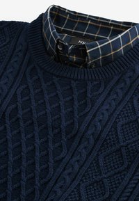 Pulover tricotat de culoare bleumarin, suprapus peste o cămașă cu colier de un plaid întunecat, având un model cu pătrate albastre, albe și maro, cu detalii cu nasturi.