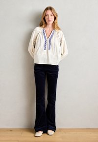 Weißes Leinenbluse mit blauen gestickten Details, kombiniert mit dunkelblauben, ausgestellten Cordhosen und beigen spitzen Schuhen.
