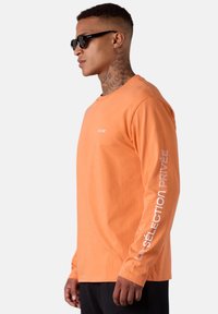 Langärmliges oranges Baumwoll-T-Shirt mit Rundhalsausschnitt. Mit weißem Text auf dem linken Ärmel und einem kleinen Logo auf der Vorderseite.