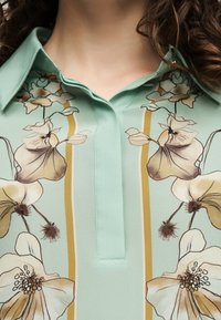 Gros plan d’une chemise vert clair avec un col et des rayures verticales jaunes, avec de grands imprimés floraux beige et marron.