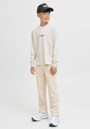 Chemise à manches longues crème avec logo noir, associée à un pantalon de jogging beige et des baskets blanches avec des accents gris ; design de tenue décontractée.