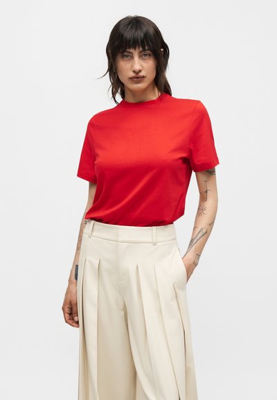 Γυναίκα με σκούρα μαλλιά φοράει ένα κόκκινο κοντομάνικο πουκάμισο πασπαλισμένο σε μπεζ πλισέ φαρδιά παντελόνια ψηλής μέσης, στέκεται με το ένα χέρι στην τσέπη.