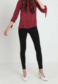 Leggings neri con vestibilità slim, dotati di strisce laterali multicolori. Abbinati a una blusa rosso scuro e scarpe con tacco bianco a blocco.