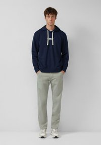 Un giovane uomo è in piedi contro uno sfondo neutro, indossando una felpa blu scuro con cordini bianchi, pantaloni grigi chiari e sneakers bianche.