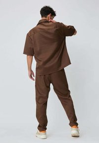 Ensemble marron deux pièces comprenant une chemise à manches courtes à coupe décontractée et un pantalon ample, tous deux en tissu texturé, associés à des baskets blanches.