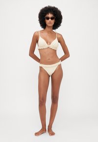 Beiges Bikini-Set mit Rüschendetails; Triangel-Bralette-Top mit verstellbaren Trägern, passende tief sitzende Unterteile. Modell steht barfuß und trägt Sonnenbrillen.