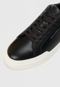 Musta nahkakengät, joissa on pyöristetty kärki, tummat kengännyörit ja valkoinen kumipohja. Sivussa on huomaamaton brändäys. Pintamateriaali on tasainen.