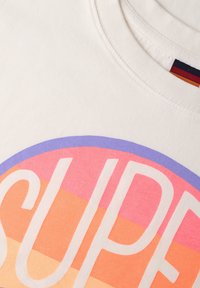 Camiseta blanca de algodón con gran texto "SUPER" en blanco bold, situado sobre un fondo circular degradado de rosa, naranja y púrpura.
