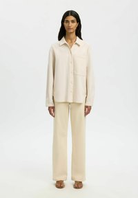 Femme portant une chemise crème ample à boutons, un pantalon large assorti et des sandales marron, debout devant un fond blanc uni.