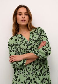 Žalia leopardų rašto marškinėliai su V formos iškirpte, ilgomis rankovėmis su sukeltomis rankovių detalėmis ir sagų puošimu priekyje. Minkšta audinio tekstūra.
