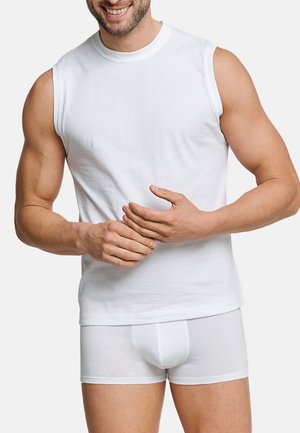 Smiling man wearing a white sleeveless shirt and white boxer briefs, with hands clasped at waist against a plain background.