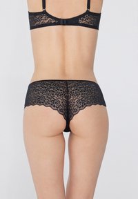 Černé krajkové prádlo sestávající z krajkového topu a kalhotek se zvlněným lemem a květinovým vzorem. Střih je krátký s nízkým pasem, což zdůrazňuje složité detaily.