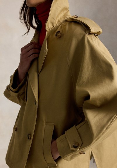 Άτομο που φοράει μπεζ καμπαρντίνα με κουμπιά πάνω από κόκκινο πουλόβερ με ζιβάγκο, με το χέρι να ρυθμίζει το παλτό κοντά στον γιακά.