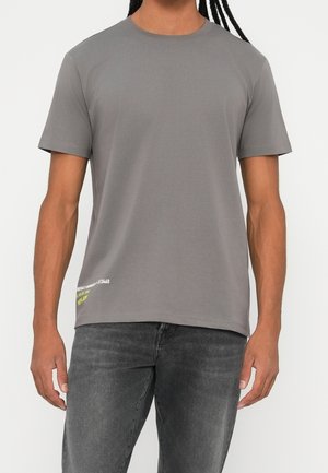Homme portant un t-shirt gris uni à manches courtes et un jean noir délavé, debout devant un fond blanc.