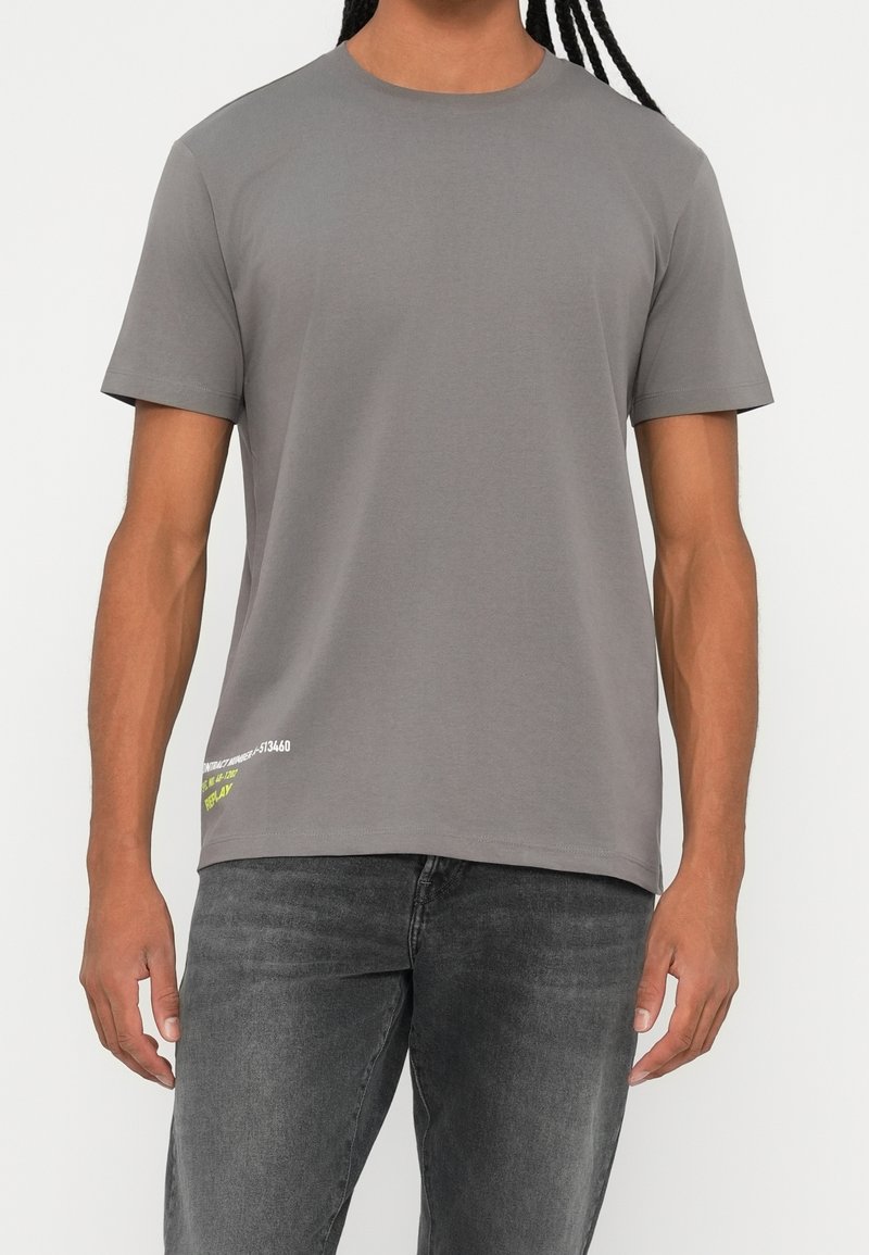 Homme portant un t-shirt gris uni à manches courtes et un jean noir délavé, debout devant un fond blanc.