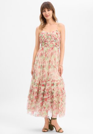 Vrouw draagt een mouwloze, bloemenpatroon enkellange jurk met spaghettibandjes en olijfgroene sandalen met blokhak op een witte achtergrond.