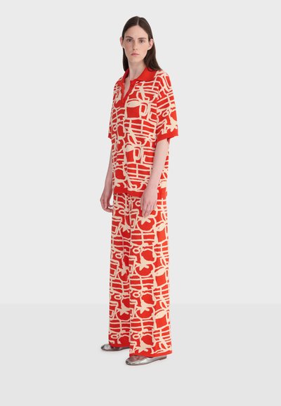 Femme debout portant un haut à manches courtes à motifs abstraits rouges et beiges assorti avec col et pantalon à jambes larges, chaussures argentées.