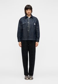 Τζιν μπουφάν με δύο μπροστινές τσέπες, σκούρο μπλε χρώμα, σετ με ριγέ πουκάμισο και μαύρο παντελόνι· ολοκληρώνεται με γκρι αθλητικά παπούτσια.