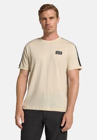 Ljusbeige t-shirt med rund hals, korta ärmar och svarta sidpaneler. Har en liten EA7-logotyp på bröstet. Mjuk bomullsmaterial.