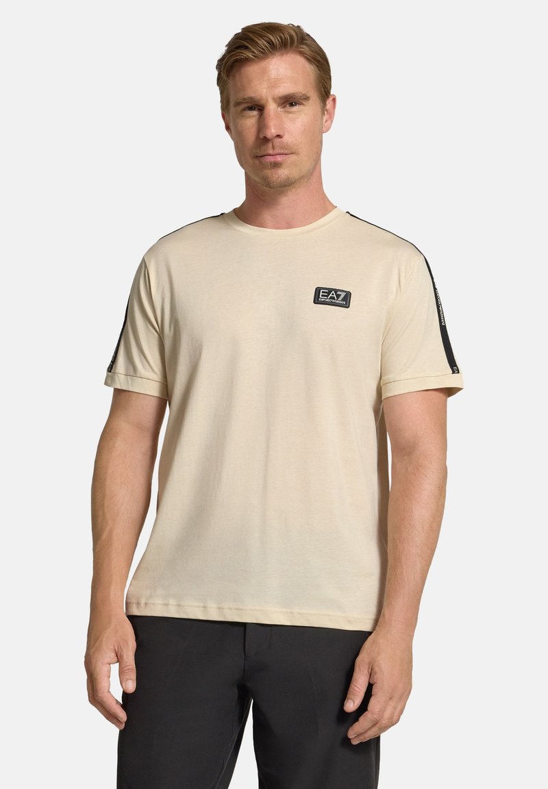 Ljusbeige t-shirt med rund hals, korta ärmar och svarta sidpaneler. Har en liten EA7-logotyp på bröstet. Mjuk bomullsmaterial.