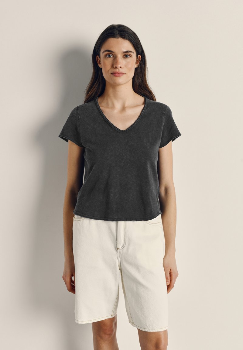 Ženska z dolgimi temnimi lasmi nosi zbledelo črno majico z V-izrezom in široke bele kratke hlače do kolen, stoji ob preprosti bež steni.