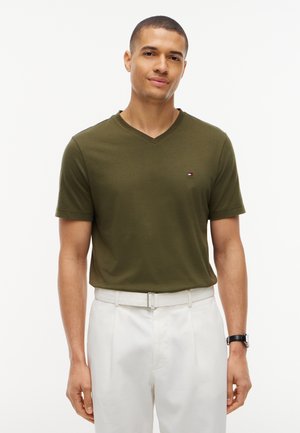 Man die een olijfgroene V-hals T-shirt en een witte broek draagt, staat voor een effen witte achtergrond.