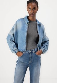 Chaqueta de denim en azul claro con un diseño cropped, puños elásticos y cierre de cremallera. Combinada con una camiseta gris y jeans azules.