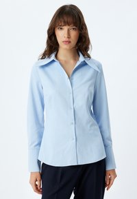 Camisa abotonada azul claro con cuello extendido, con mangas largas y cintura ajustada, hecha de una tela suave, diseñada para un aspecto pulido.