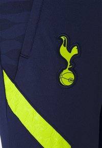 Pantalones cortos deportivos de color navy con un tejido texturizado, una franja diagonal amarilla brillante y un logo de club bordado en amarillo.