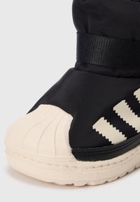 Baskets noires et blanches avec un embout en caoutchouc ribbed blanc cassé et trois rayures blanches en zigzag sur le côté, le tout sur un fond uni.