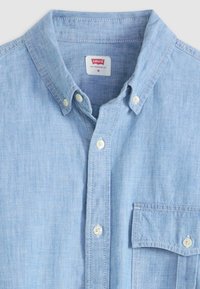 Lyseblå skjorte med knapper lavet af chambray-stof. Har en button-down krave, brystlomme foran og hvide knapper.