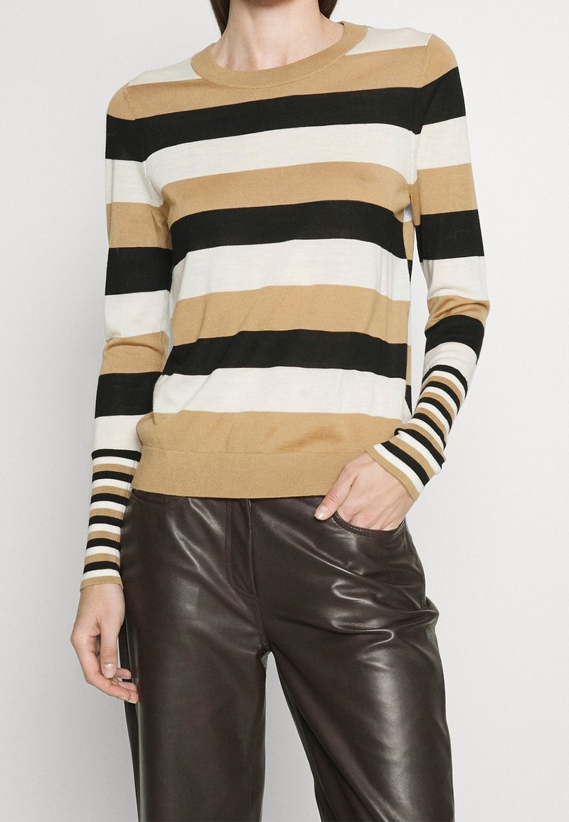 Femme portant un pull à manches longues rayé beige, noir et blanc avec un pantalon en cuir marron, une main dans la poche, sur un fond uni.