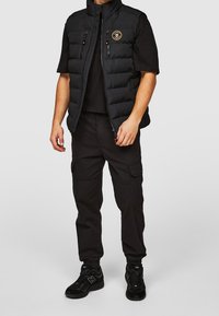 Czarna pikowana kamizelka z wysokim kołnierzem, dwiema kieszeniami na zamek i naszywką z logo, noszona na czarnej koszuli, w połączeniu z czarnymi spodniami typu cargo i sneakersami.