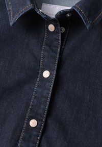Chemise en denim bleu foncé en gros plan montrant le col, les boutons-pression et les surpiqûres contrastées le long de la patte de boutonnage.
