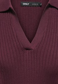 Gros plan d'un col en maille côtelée violet foncé avec encolure en V et une étiquette noire de vêtement indiquant ONLY OUR STORY taille S.