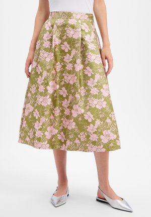 Frau trägt einen olivgrünen Midirock mit pinkem Blumenmuster, kombiniert mit weißem Oberteil und silbernen spitz zulaufenden Slingback-Ballerinas.