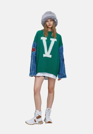Ung kvinde iført en oversize grøn sweater med stort hvidt V, denimærmer med rosenpatch, grå pels hat, hvide sneakers og kort hvid nederdel.