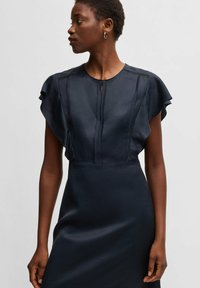 Marinblå klänning med korta fladderärmar, som har en rund halsringning och en detalj i form av en nyckelhål framtill. Slät tyg med subtil lyster och en figursydd silhuett.