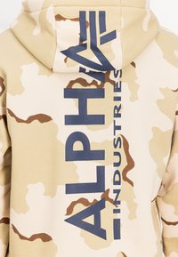 Hoodie camouflage beige avec le texte "ALPHA INDUSTRIES" en bleu foncé bien en vue au dos, doté d'une texture douce et d'une capuche réglable.