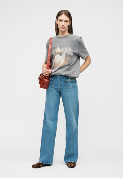 Γυναίκα στέκεται φορώντας γκρι μπλούζα με στάμπα δύο αλόγων, μπλε τζιν παντελόνι με φαρδιά μπατζάκια, παπούτσια με λεοπάρ μοτίβο και κόκκινη τσάντα ώμου με μεταλλικά ανοίγματα.