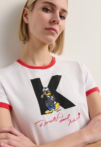 Balta marškinėliai su raudonais kraštais, kuriuose yra išsiuvinėtas Donaldas Dakas ir ryški juoda raidė "K" su parašu raudonai apačioje.