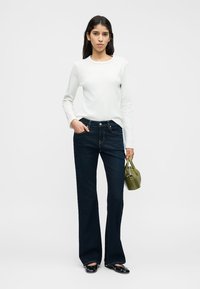 Λευκό μακρυμάνικο τοπ, σκούρα μπλε τζιν με καμπάνα, μαύρα παπούτσια. Κρατάει μια μικρή τσάντα χειρός σε ελιά πράσινο χρώμα. Απλός, εφαρμοστός σχεδιασμός με ελάχιστες λεπτομέρειες.