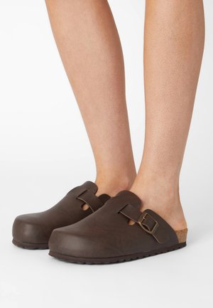 Brown clogs with a closed toe and side cutouts. Made of smooth material, featuring a metal buckle and a textured cork sole.