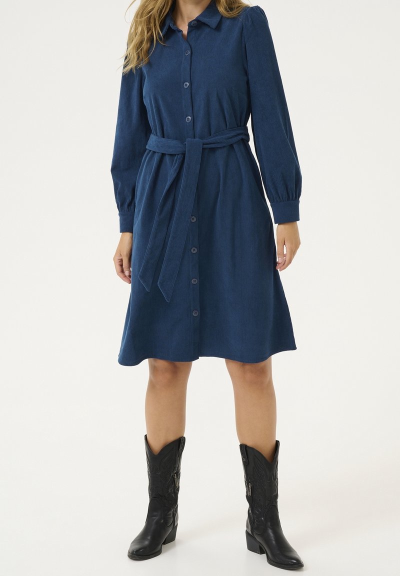 Une femme porte une robe bleu marine boutonnée jusqu'aux genoux, à manches longues et avec un lien à la taille, associée à des bottes de cow-boy noires brodées.