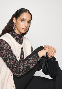 Bolsa preta com um acabamento texturizado, blusa com padrão floral em tons escuros, colete de malha rosa pálido e calças pretas justas.