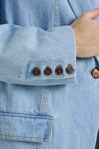 Manche d'une veste en denim bleu clair avec quatre boutons marron à l'apparence légèrement brillante et des accents dorés ; détails de couture visibles sur le tissu.