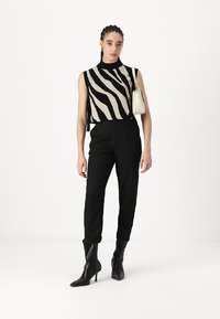 Haut sans manches noir et beige à rayures, col montant. Pantalon noir à rayures fines, jambes courtes. Bottes à talons noires. Petit sac blanc. Fond uni.