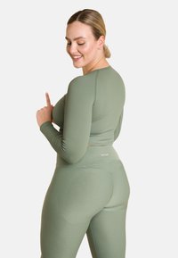 Olivenfarbener Sportanzug bestehend aus einem gerippten Langarmoberteil und hochgeschnittenen Leggings, beide mit einer figurbetonten Passform und weichem Material.