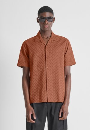 Hombre con gafas de sol oscuras y camisa marrón de manga corta con botones, con un patrón de ojales en forma de chevron, y pantalones negros, de pie recto.