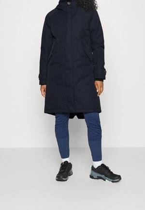 Persona con un abrigo azul marino hasta la rodilla, pantalones ajustados azules, calcetines blancos y zapatos deportivos negros, de pie frente a un fondo liso.